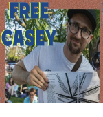 Free Casey Image of white person in white tshirt with black hat and black glasses holding prisoner solidarity zines.